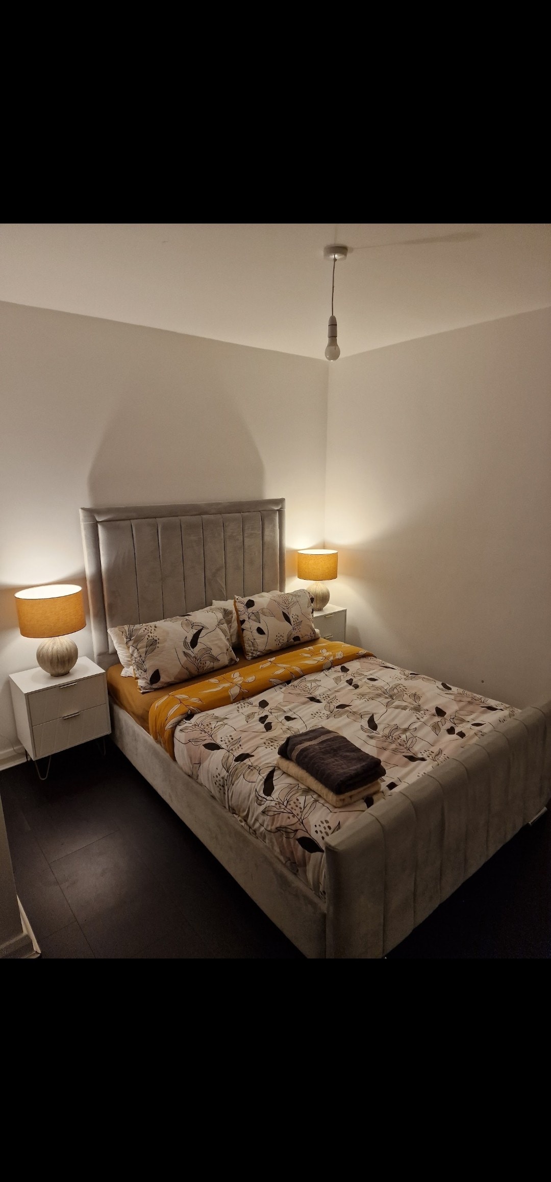 A serene bedroom features a plush bed with decorative pillows and a coordinated blanket. Soft lighting from two bedside lamps casts a warm glow, while a sleek nightstand holds a textured lamp. Dark flooring contrasts with the light walls, creating a calm environment.