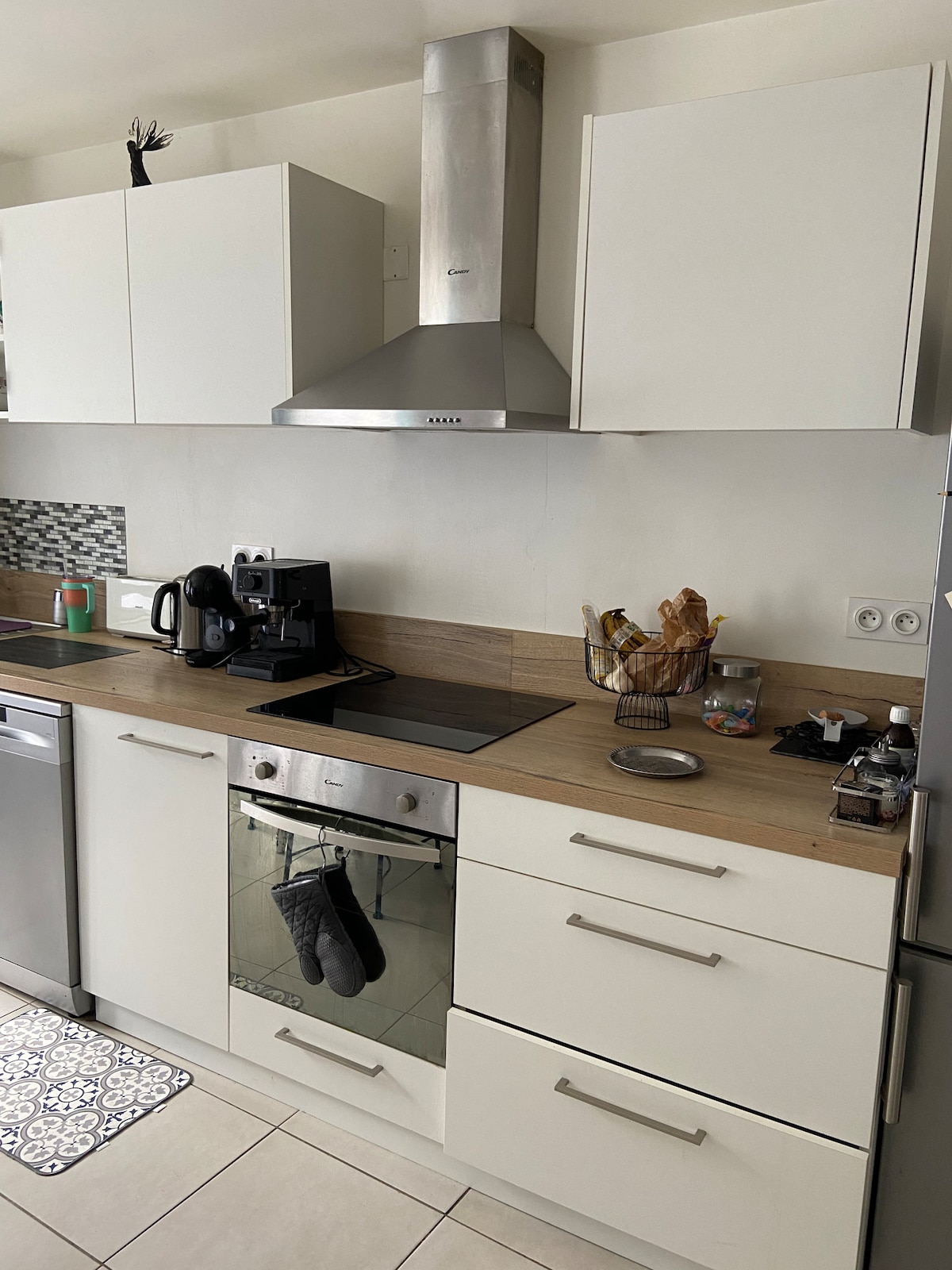 A modern kitchen setup features sleek white cabinetry and a wooden countertop. Essential appliances include a built-in stovetop and a kettle. A coffee maker and various utensils are arranged neatly, while two dark oven mitts hang on the front of the oven. Natural light brightens the space.