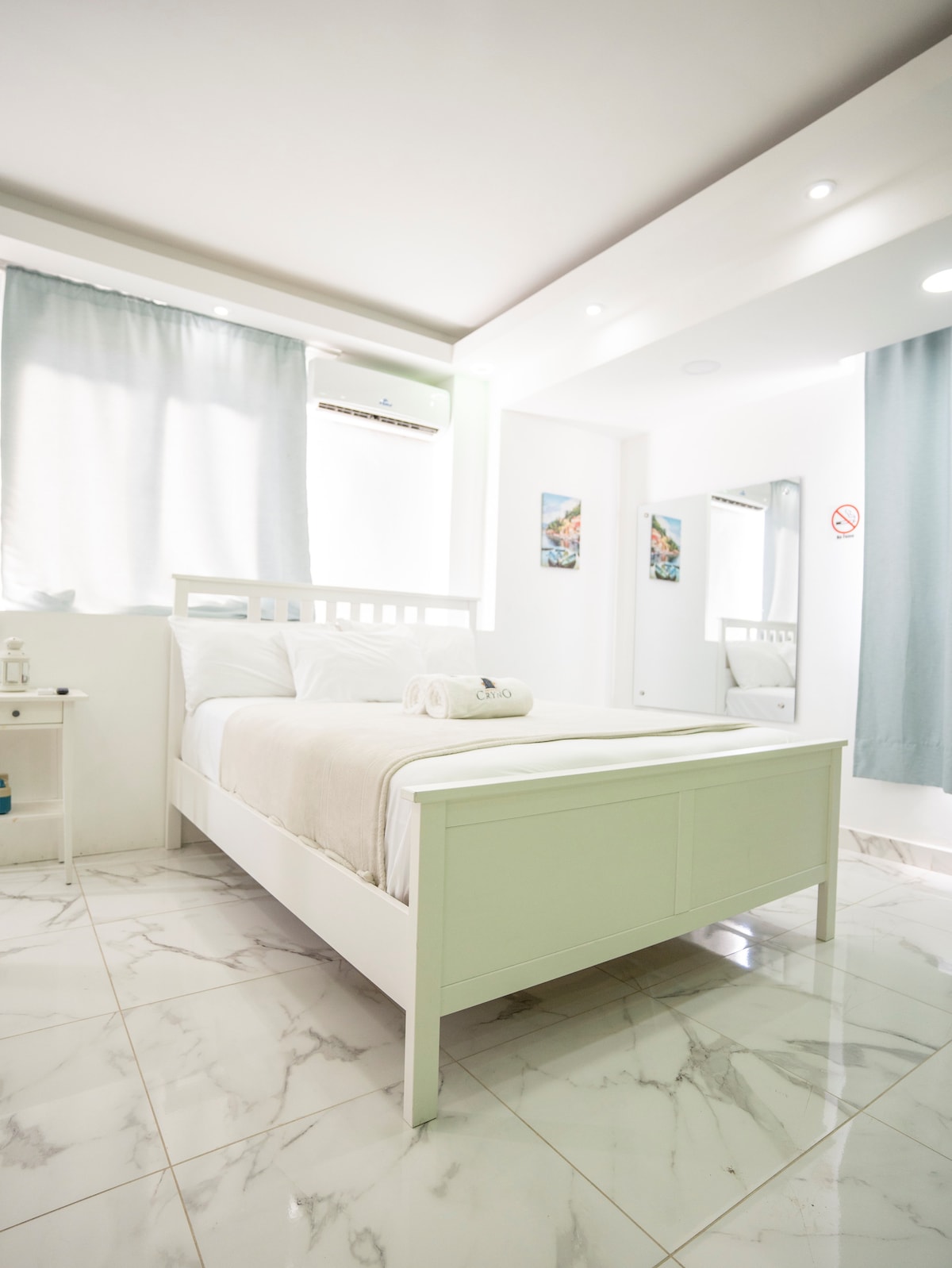 A well-lit bedroom showcases a spacious white bed adorned with soft linens. Two windows dressed in light curtains allow natural light to fill the room. Reflections can be seen in the wall mirror, complementing the minimalist decor highlighted by marble flooring.