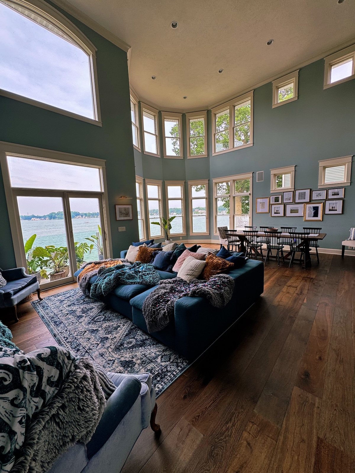 An open-concept living area features large windows providing panoramic views of the lake. A plush dark sofa is adorned with cozy throw pillows, complemented by a patterned area rug. A dining table with black chairs is positioned nearby, enhancing the welcoming atmosphere.