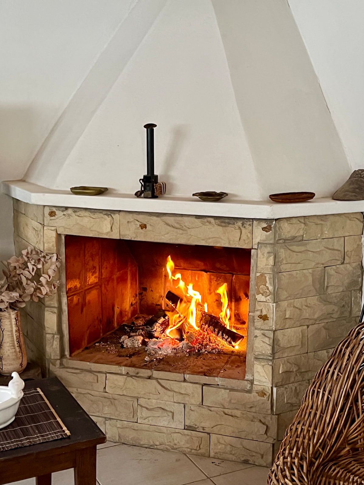 A stone fireplace is featured in the living space, with flames brightly burning within the hearth. The fireplace is surrounded by a simple white mantel and decorated with a few small bowls. Nearby, a woven chair is positioned for comfort.