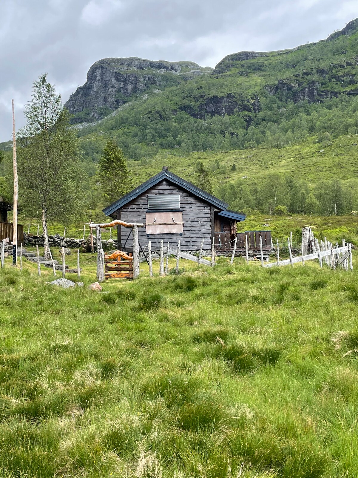 A rustic wooden cabin is viewed from a grassy foreground, surrounded by lush greenery and distant mountains. The exterior features boarded windows and a sloped roof, with a split-rail fence enclosing the area. Natural light casts a soft glow on the landscape.