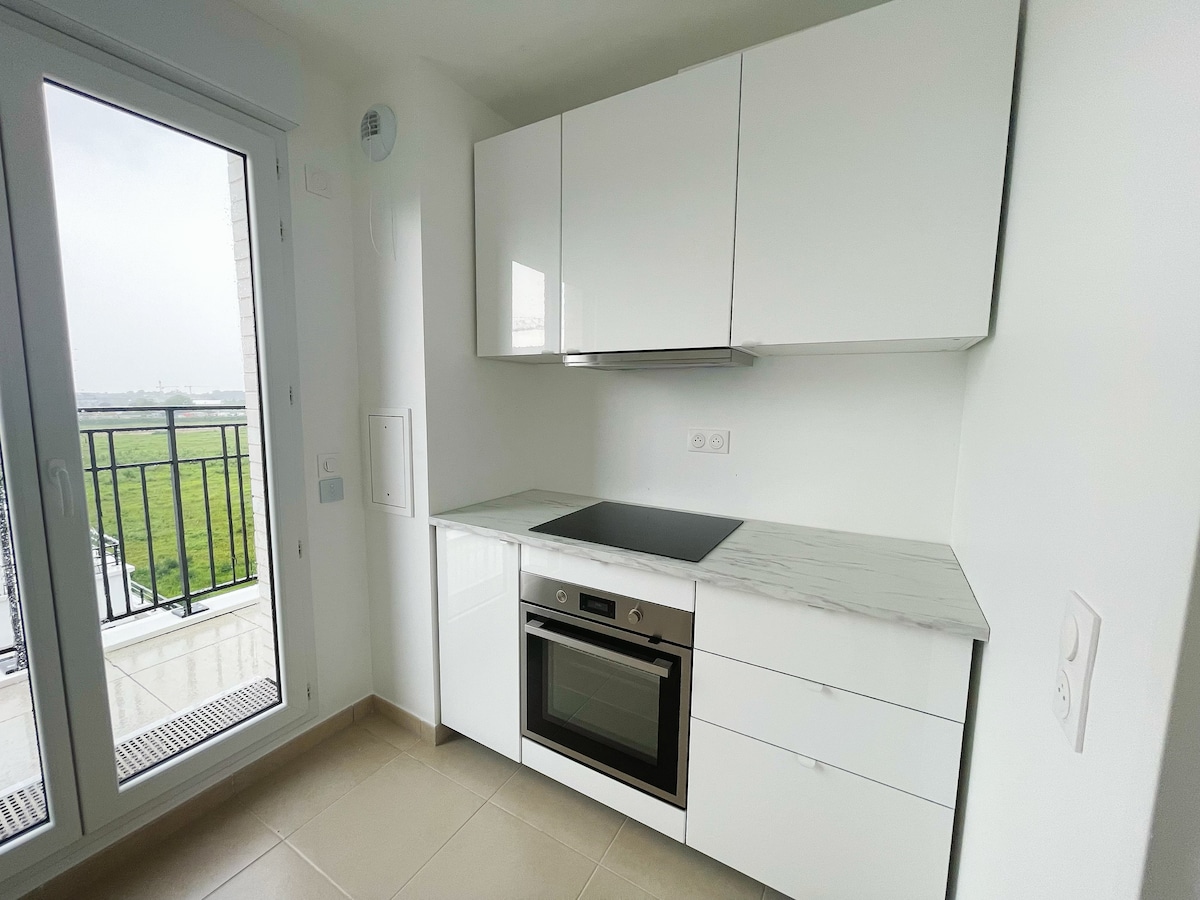 A bright, modern kitchen features sleek white cabinetry and a marble countertop. An integrated oven and cooktop are positioned beneath the counter. A glass sliding door provides access to a small balcony, offering a view of the greenery outside.