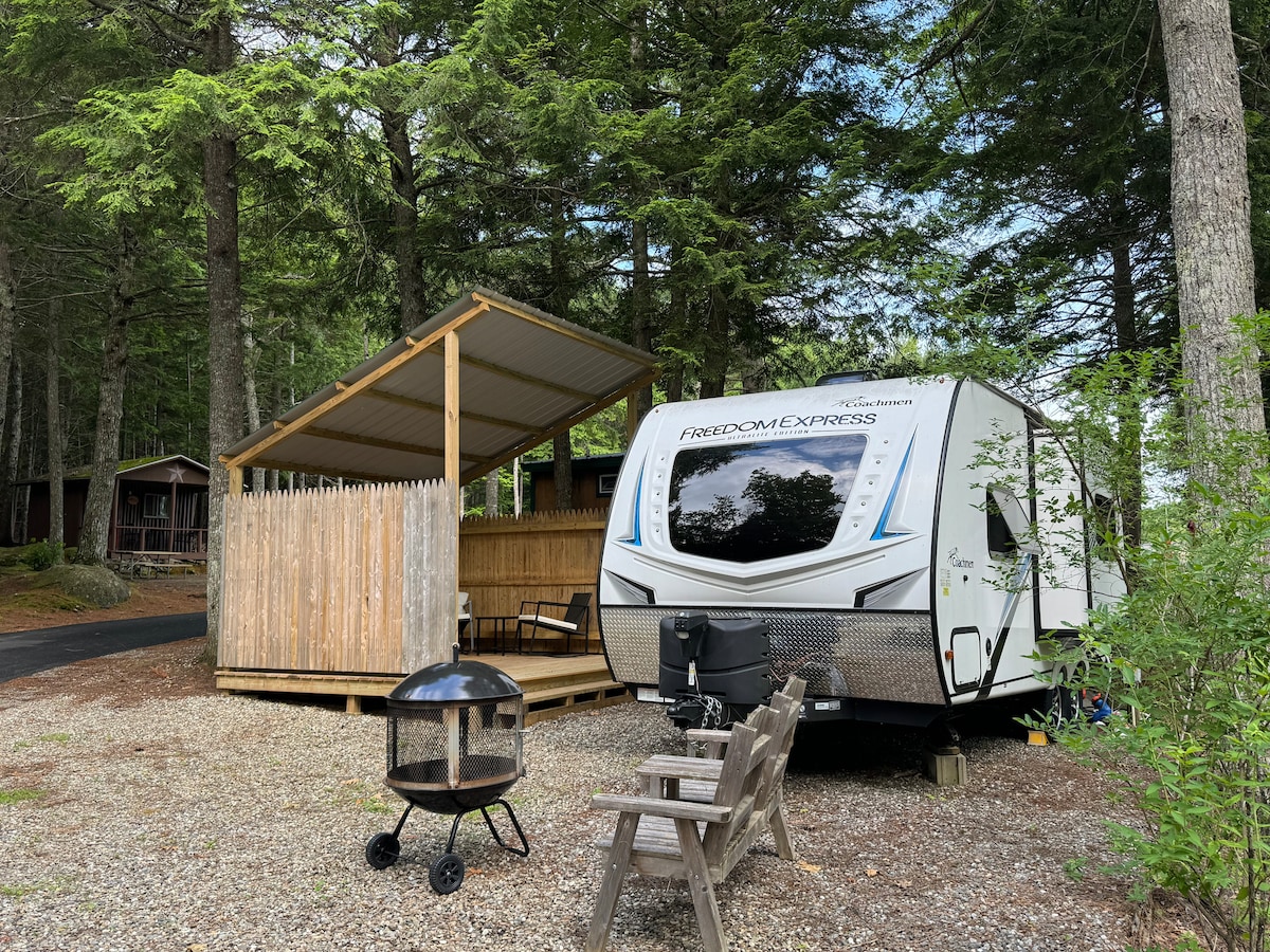 A compact RV is parked on a gravel area, surrounded by tall trees. A wooden gazebo provides shelter with open seating nearby. A small fire pit is positioned on the ground, complementing the outdoor space for relaxation.