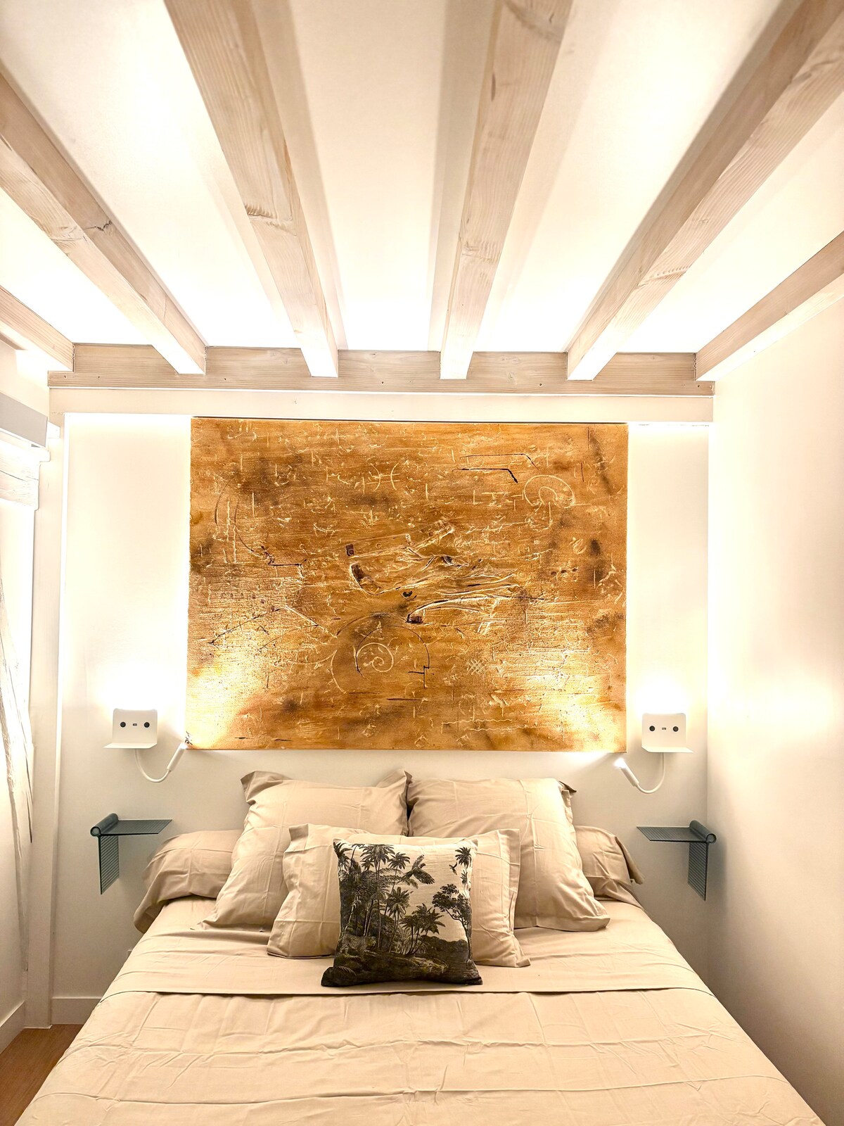 A serene sleeping area features a spacious bed with soft beige linens and an accent pillow adorned with a tropical design. The wall above is highlighted by a warm, textured artwork, complemented by integrated lighting and wooden beams overhead, creating a calming ambiance.