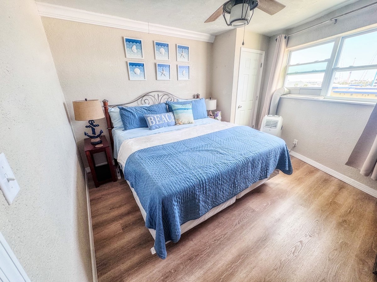 استرخ في غرفة نوم ساحلية هادئة مع سرير بحجم كينج، وأغطية سرير زرقاء ناعمة، ومصابيح بجانب السرير، ومروحة سقف لمزيد من الراحة. تخلق الأرضيات الخشبية الخفيفة والنافذة المشمسة ملاذًا هادئًا بعد يوم على الشاطئ.
