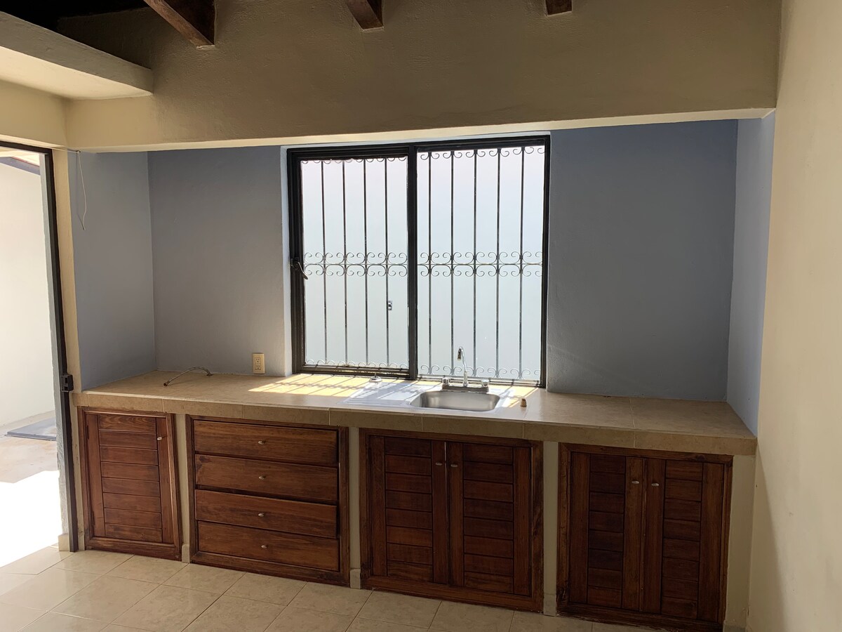 A functional kitchenette is presented with wooden cabinetry and a countertop. A small sink is positioned beneath a window with security bars, allowing natural light to illuminate the space. The neutral walls are complemented by tiled flooring.