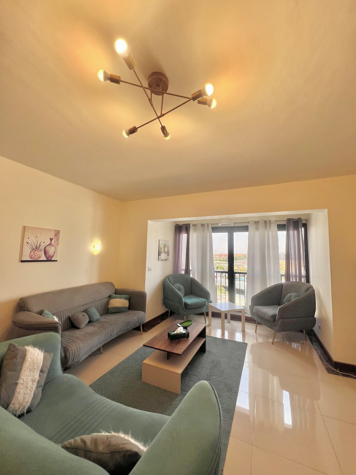 A welcoming living area is presented, featuring two plush armchairs and a comfortable sofa. A coffee table sits centrally atop a soft rug, and large windows with sheer curtains invite natural light, offering views of the surrounding area. Simple wall art enhances the ambiance.