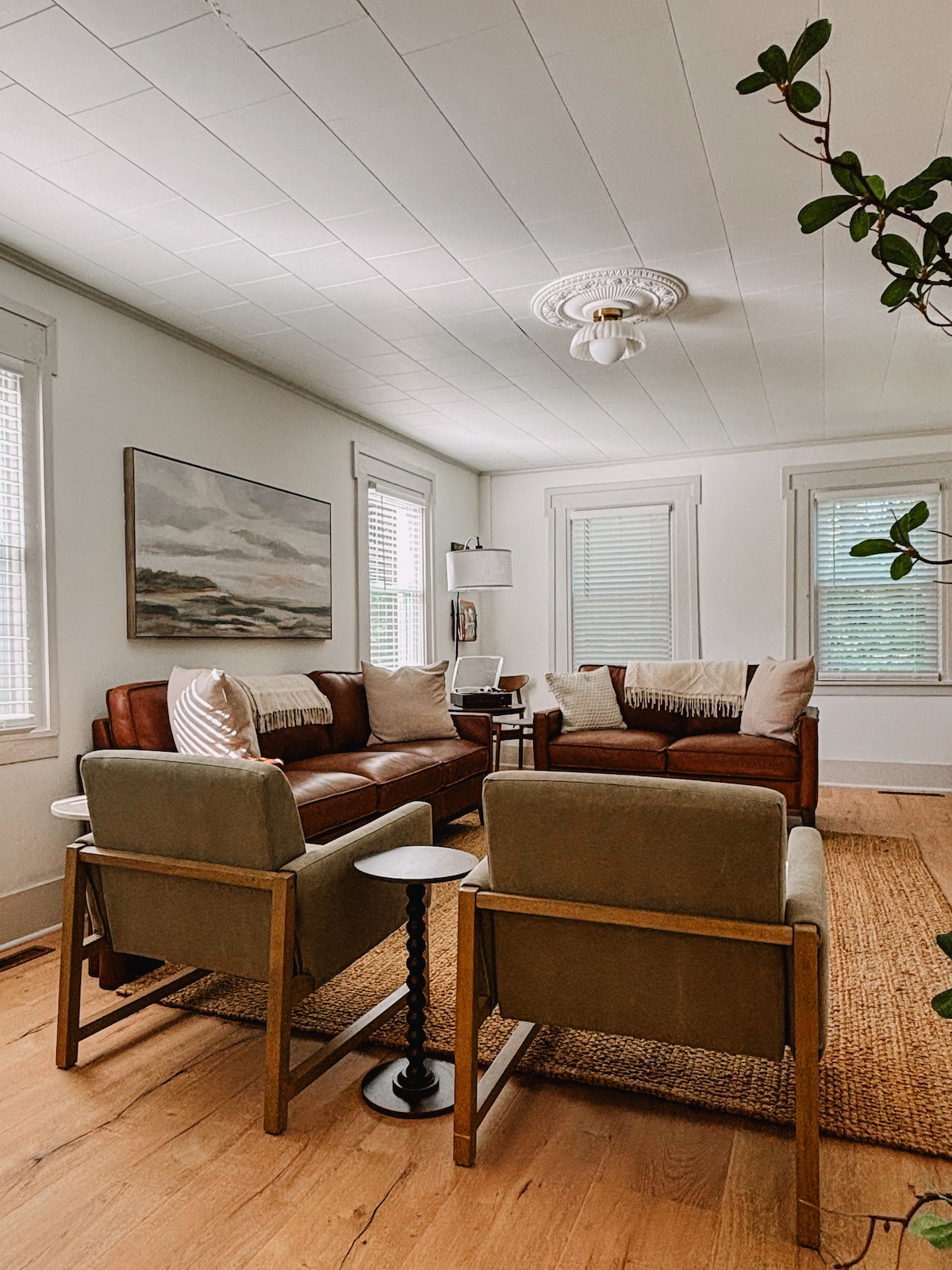 A comfortable living area is showcased, featuring a pair of brown leather sofas and two light gray armchairs. A small black side table stands beside the seating arrangement, while natural light filters through nearby windows with white blinds.