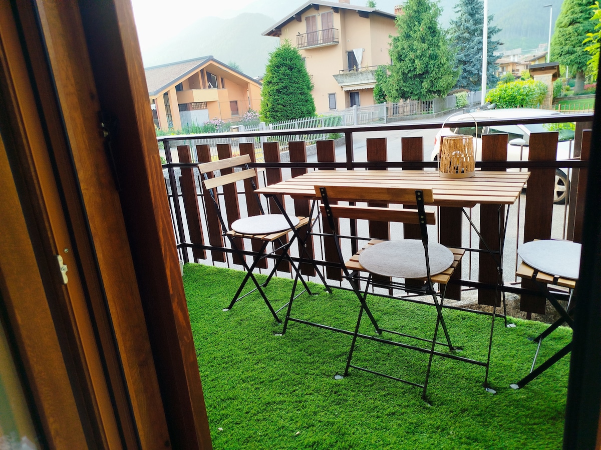 A small outdoor terrace is visible with a wooden table and two folding chairs set against a backdrop of lush green grass. The terrace is enclosed by a wooden railing, providing an inviting space for relaxation. A distant view of residential buildings and greenery can be seen.