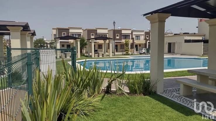 A well-maintained swimming pool is surrounded by lush greenery and neatly paved pathways. Comfortable seating areas are visible under shaded structures near the pool, providing a serene environment. The layout suggests a welcoming community space ideal for relaxation and leisure.