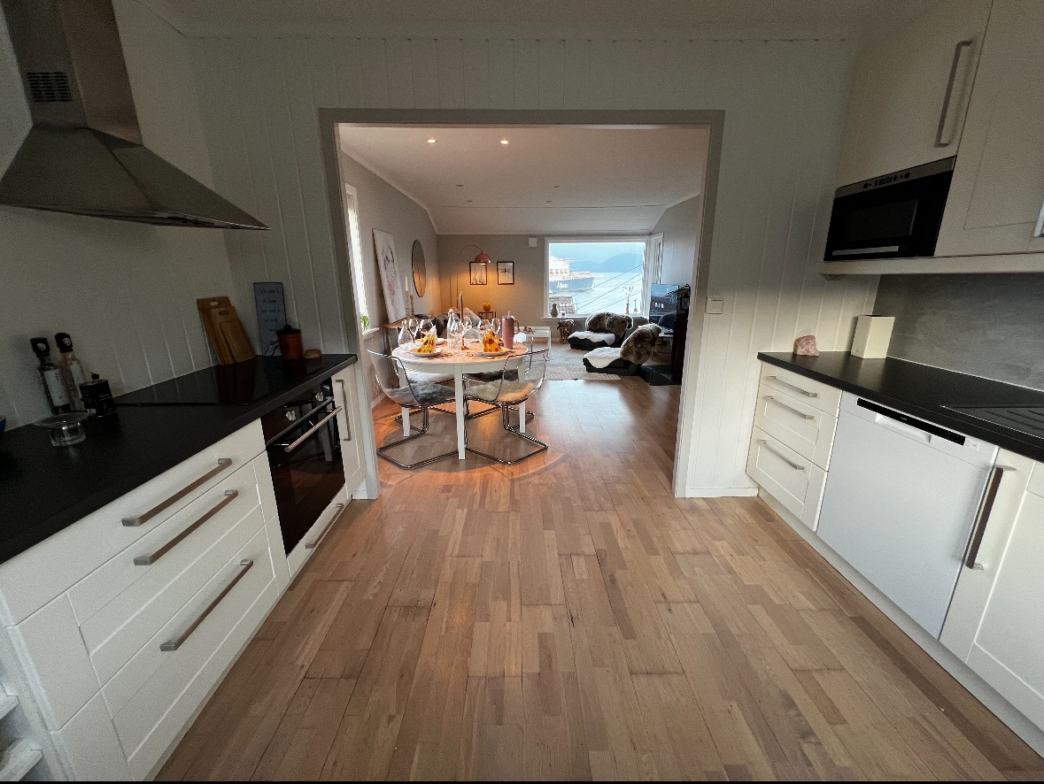 A modern kitchen is equipped with sleek white cabinets and dark countertops. The open layout leads to a dining area with a circular table set for a meal, visible through an archway. Soft lighting and a view of the fjord can be glimpsed from the living space beyond.