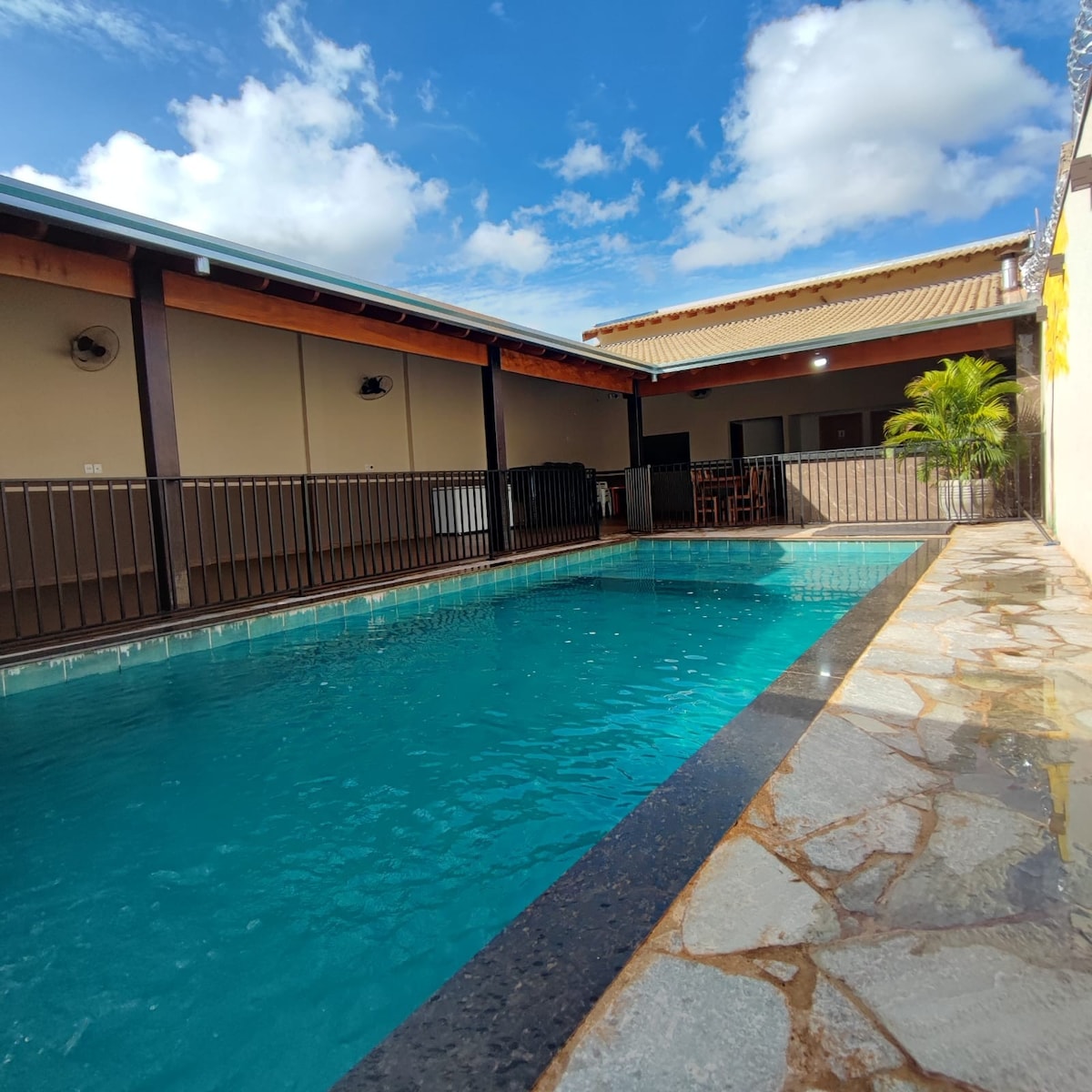 A clear blue pool, framed by natural stone tiles, reflects the bright sky above. Adjacent to the pool, a shaded area features wooden furniture, providing a place for relaxation and socializing. The overall setting is designed for comfort and leisure.