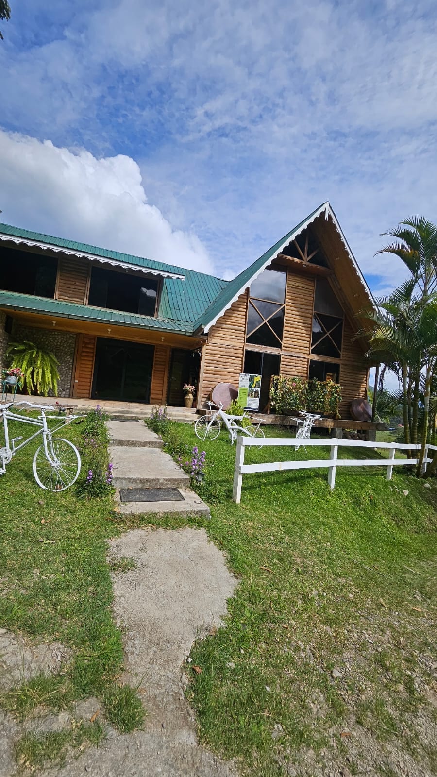 The exterior of a wooden lodge is shown, featuring a steeply pitched roof and large windows. A pathway lined with greenery leads to the entrance. Decorative bicycles are positioned along the walkway, and potted plants add color to the surrounding area.