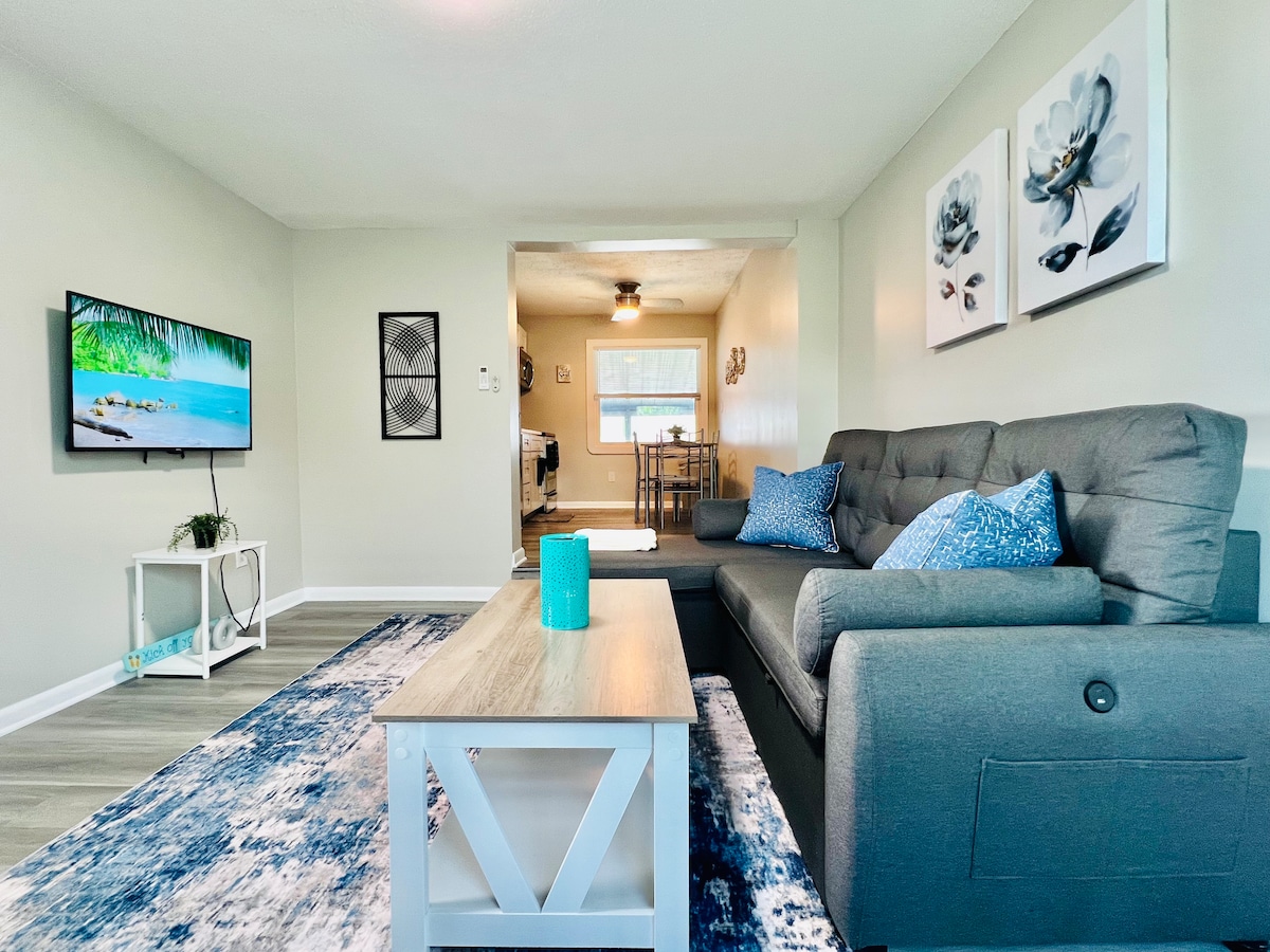 The living area is furnished with a gray sofa featuring decorative blue pillows. A coffee table is positioned on a patterned area rug, and a flat-screen TV is mounted on the wall. A doorway leads to the dining area and kitchen, with natural light flowing in from a nearby window.