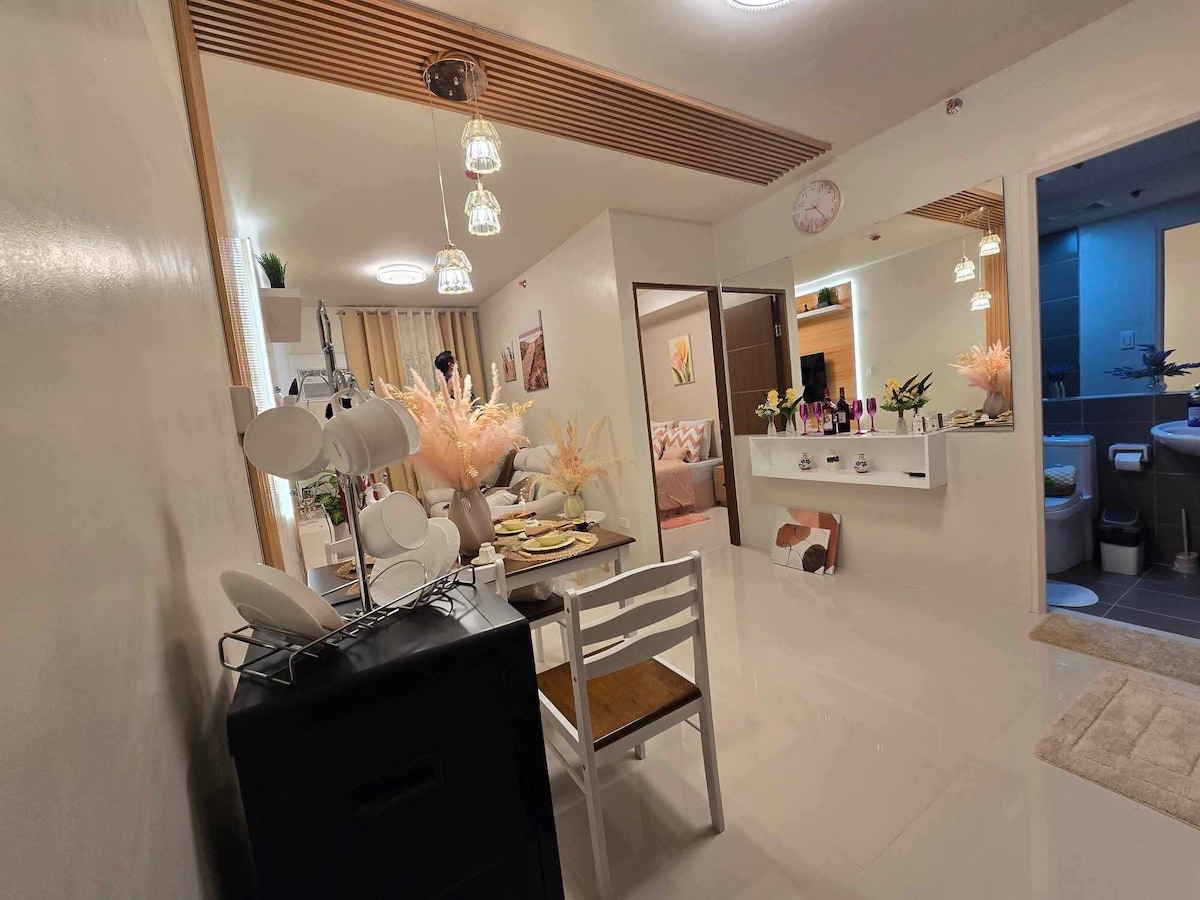 A modern dining area is visible, featuring a wooden table set for a meal, surrounded by white chairs. Soft lighting is provided by pendant lamps above. A vanity area with decorative elements is seen in the corner, while a hallway leads to a cozy living space and restroom.