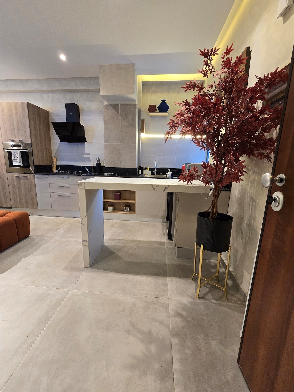 A modern kitchen area features a minimalist design with sleek cabinetry and a contrasting countertop. A decorative plant adds a touch of color next to a spacious dining table. Soft, ambient lighting highlights the neutral tones of the room, enhancing the contemporary setting.