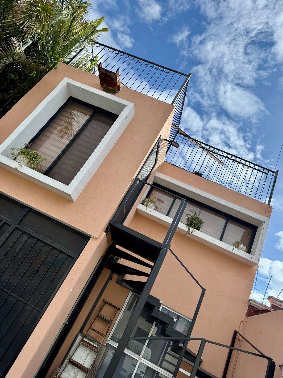 A charming two-story building is shown, featuring a soft peach exterior with contrasting white window frames. Two windows showcase potted plants. A metal staircase leads to an upper level with a balcony, surrounded by greenery, under a bright blue sky decorated with wispy clouds.
