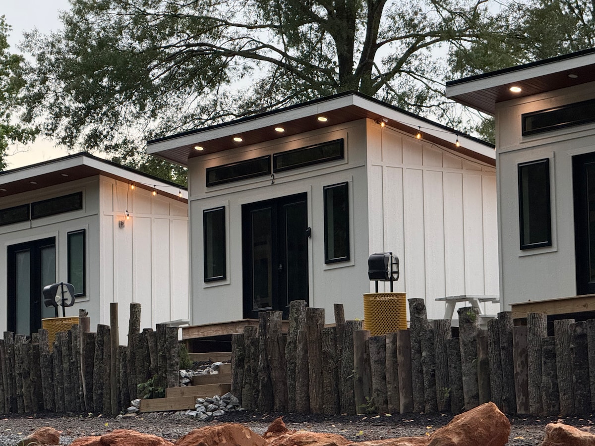 A row of modern cabins is set against a natural backdrop, featuring sleek exteriors with large windows and warm outdoor lighting. A wooden fence made of logs borders the space, enhancing the cabin's rustic charm while offering a sense of privacy and connection to nature.