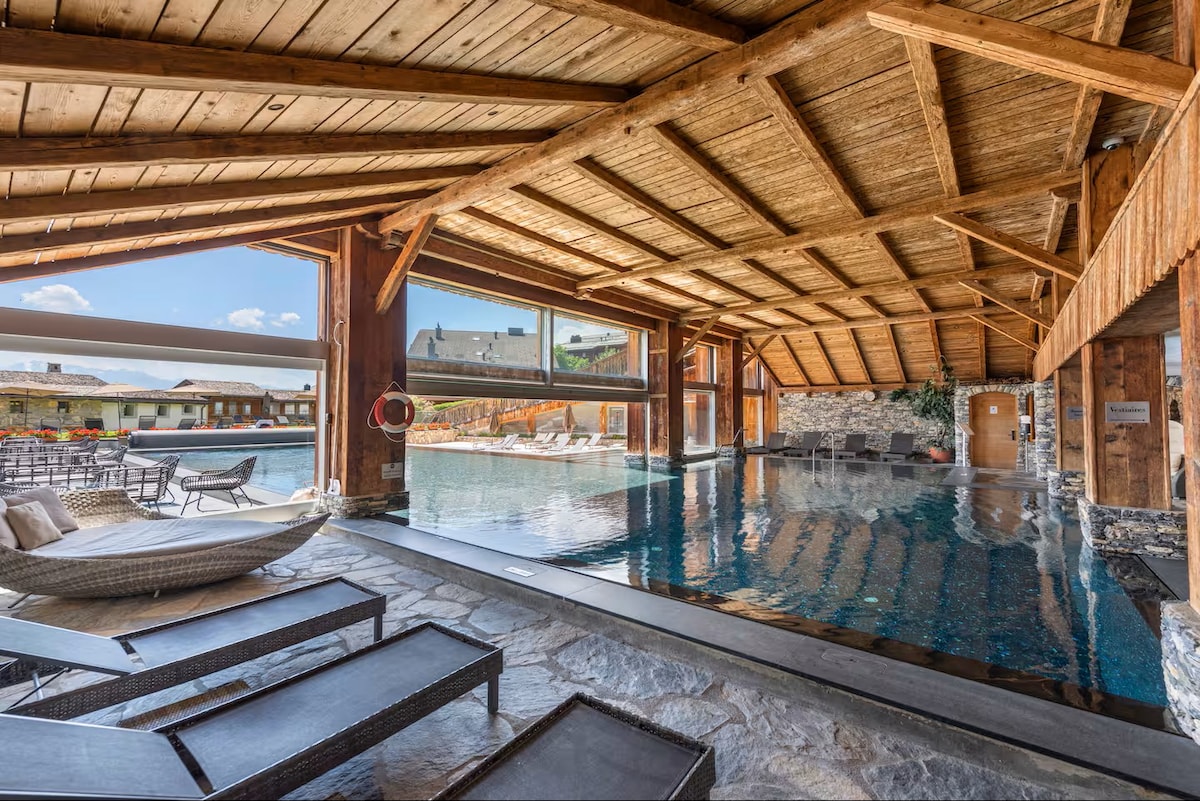 An indoor pool area is highlighted by wooden beams and stone accents. Sunlight floods the space through large glass doors, which open to an outdoor deck with lounge chairs. The inviting water reflects the surrounding architecture, enhancing the tranquil atmosphere.