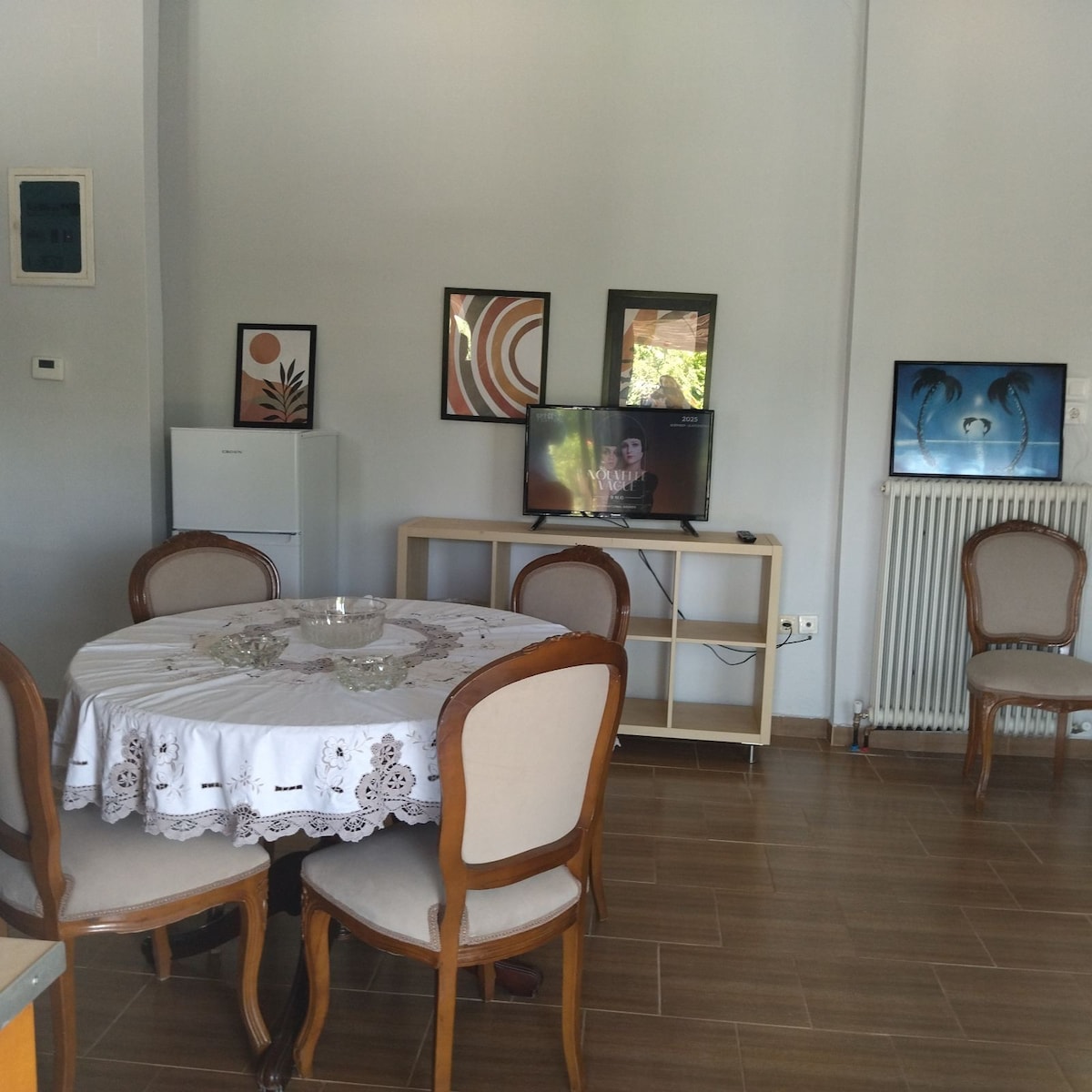 A dining area features a round table covered with a lace tablecloth, surrounded by four wooden chairs. A refrigerator is positioned near the wall, while a small media console displays a television. Wall art adds a touch of color to the space.