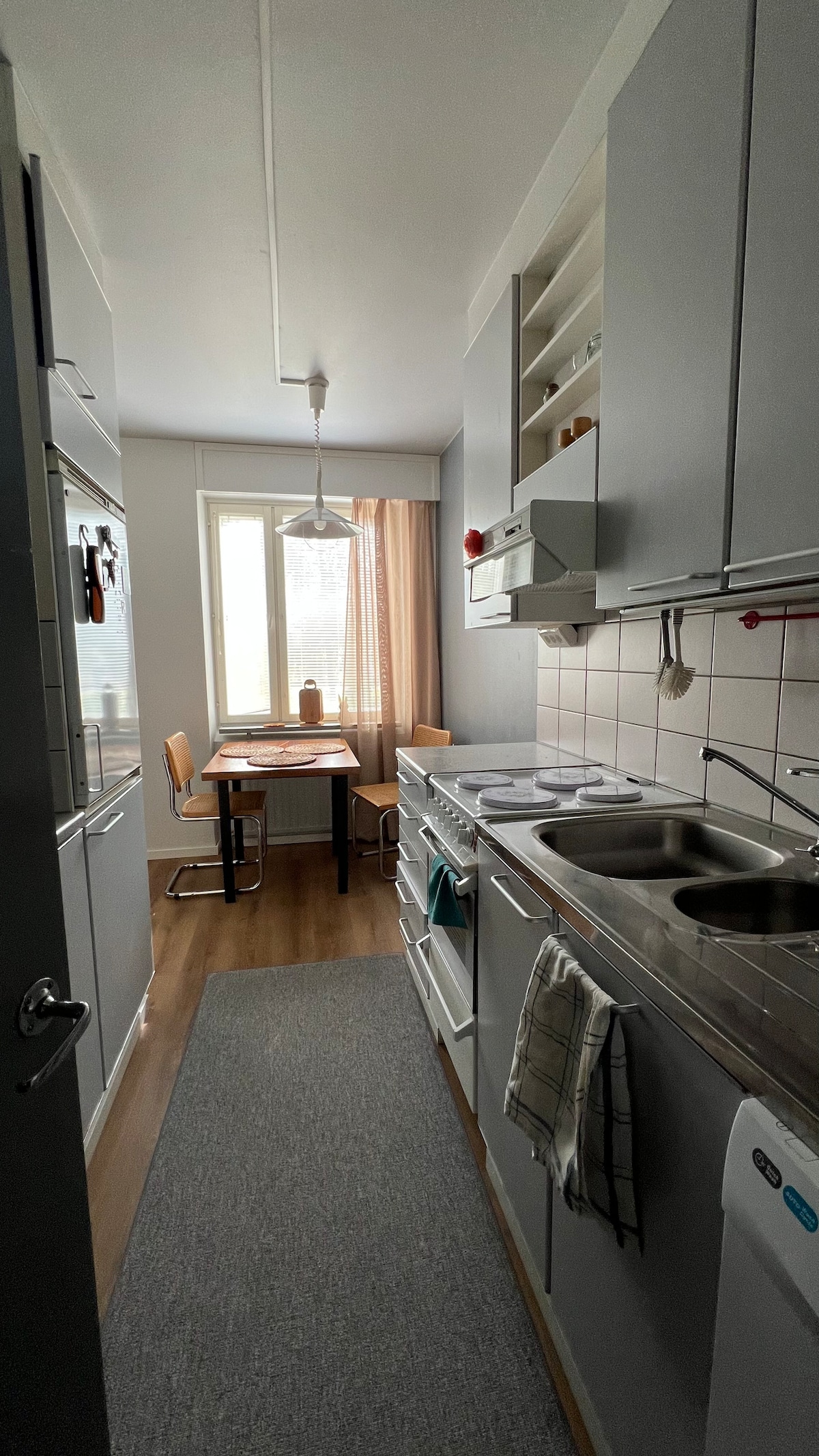 A kitchen space features modern appliances and sleek cabinetry. A wooden table with two chairs is situated near a window, providing natural light. Soft curtains frame the window, and a neutral rug runs along the floor, complementing the minimalist design.