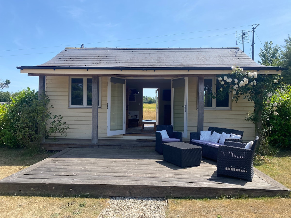A charming cabin is presented with a spacious wooden deck, featuring a comfortable outdoor seating area. Double doors open to reveal a bright interior, while lush greenery and a field frame the serene setting. A soft blue sky enhances the inviting atmosphere.