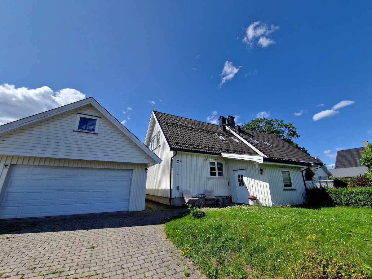A two-story house is presented alongside an attached garage. The home's exterior is painted in a light color, featuring a sloped black roof. A grassy area with wildflowers extends in front, under a clear blue sky dotted with a few clouds.