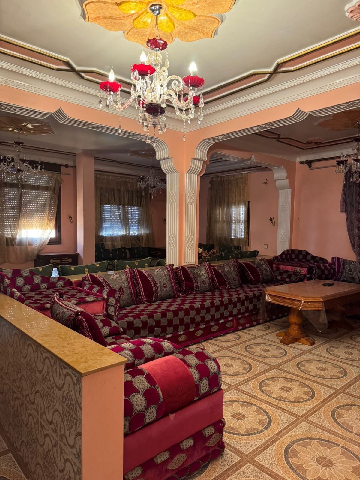 A spacious living area features plush seating in rich burgundy and patterned upholstery. Intricate ceiling moldings and a decorative chandelier add elegance. Large windows are dressed with flowing curtains, allowing natural light to brighten the space. A wooden coffee table serves as a focal point.