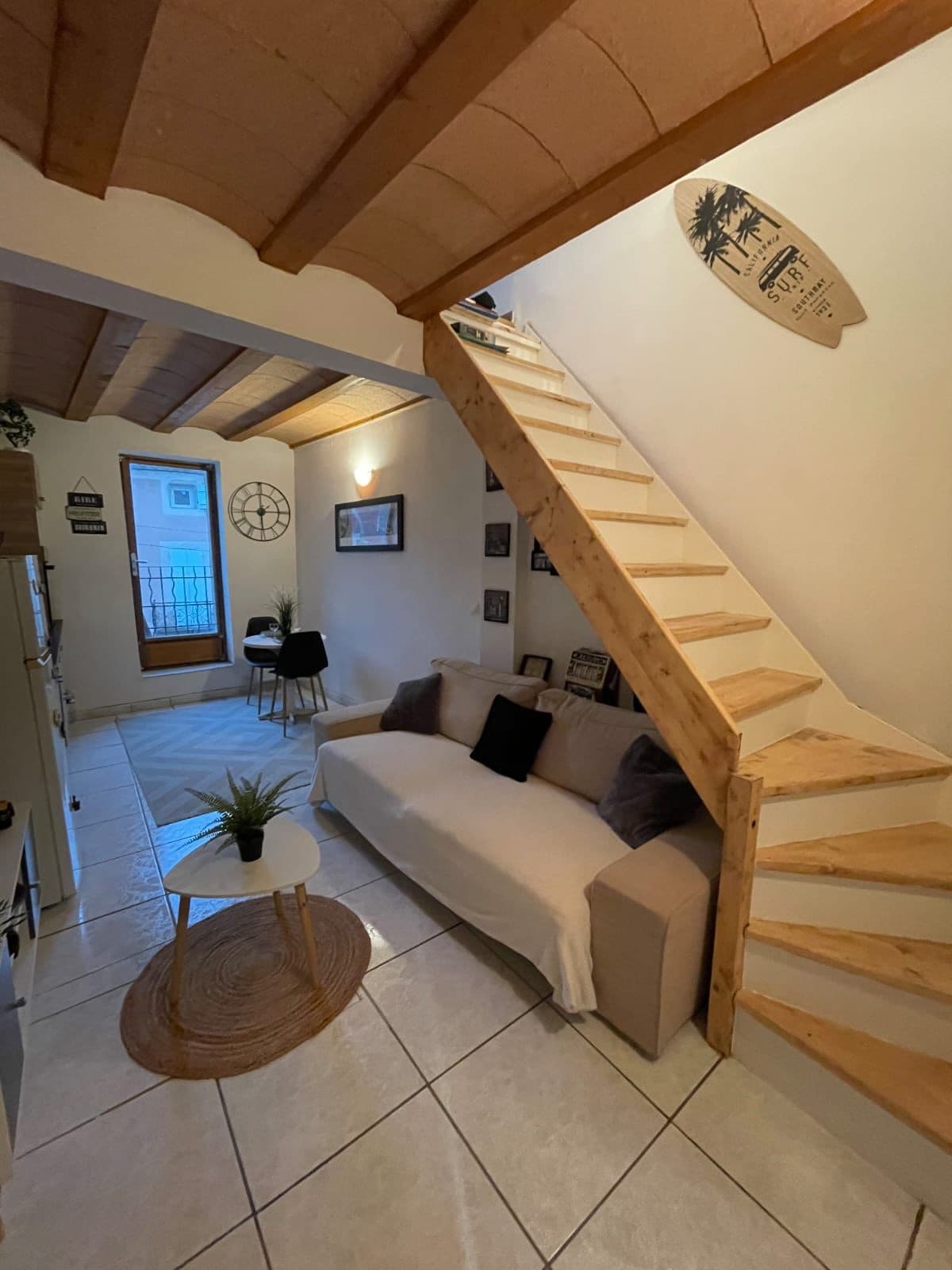The interior space features a light-colored sofa paired with dark throw pillows, positioned beside a circular coffee table. A woven rug lies under the table, while the wooden staircase leads to the upper level. Natural light enters through a window, illuminating the tiled floor.