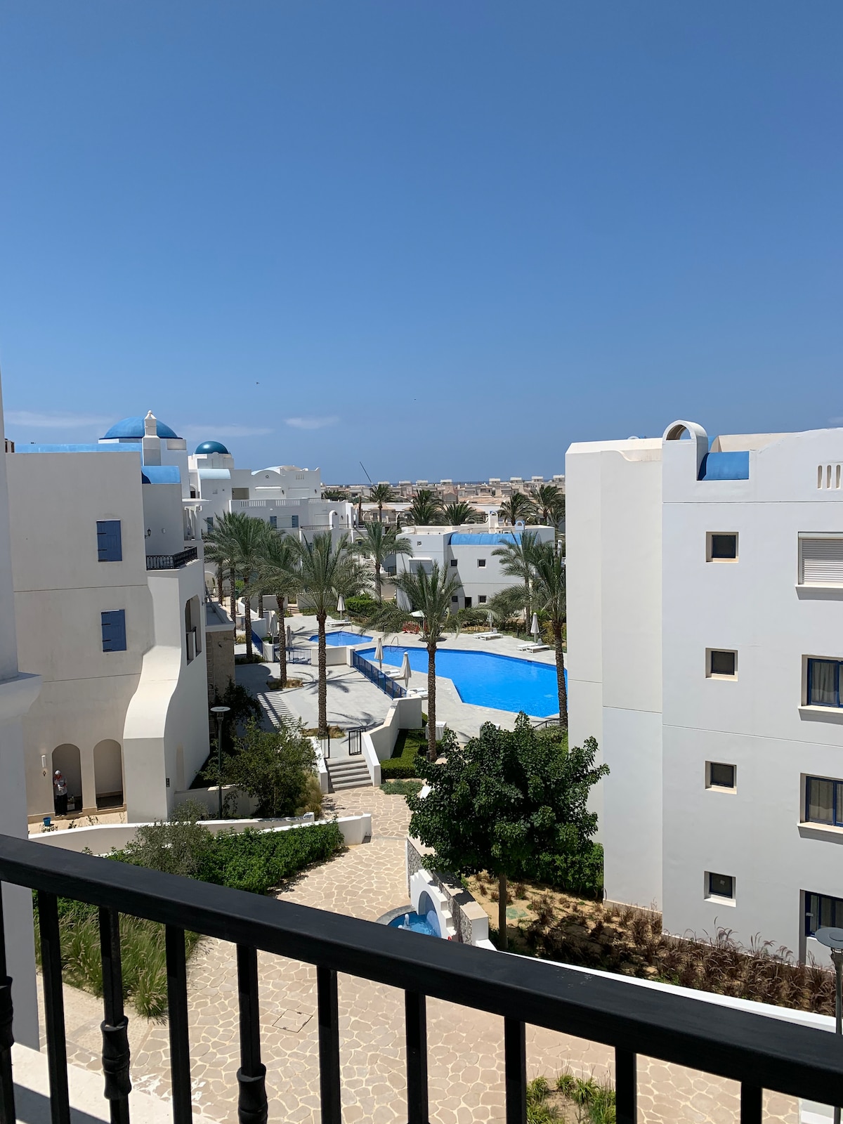 A scenic view reveals a tranquil pool nestled within the white structures of the Marassi Greek village. Palm trees line the pathways, and the bright blue sky adds to the serene ambiance. The surrounding buildings feature distinct architectural elements, enhancing the overall aesthetic.