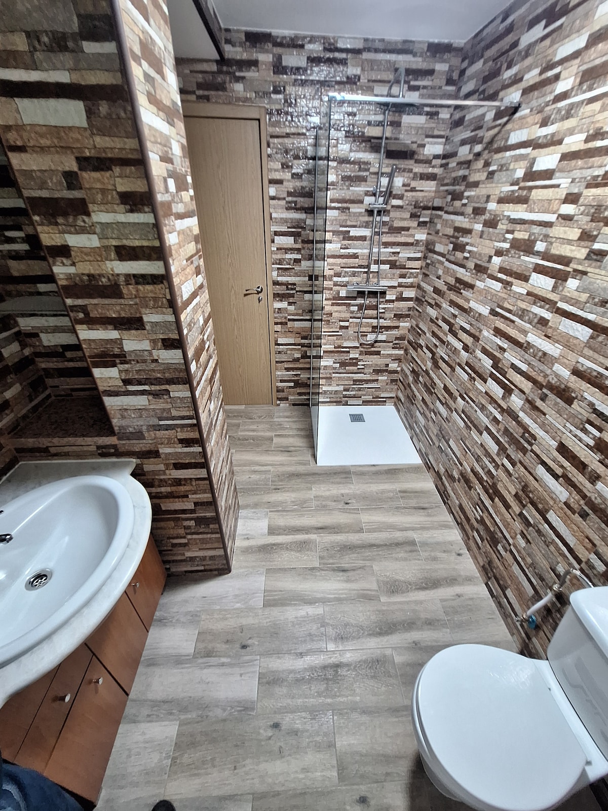 The bathroom features a spacious layout with a shower area enclosed by clear glass. A modern sink is set against a textured stone wall, complementing wooden cabinetry. Light-colored tile flooring extends throughout the space, creating a clean and neutral environment.