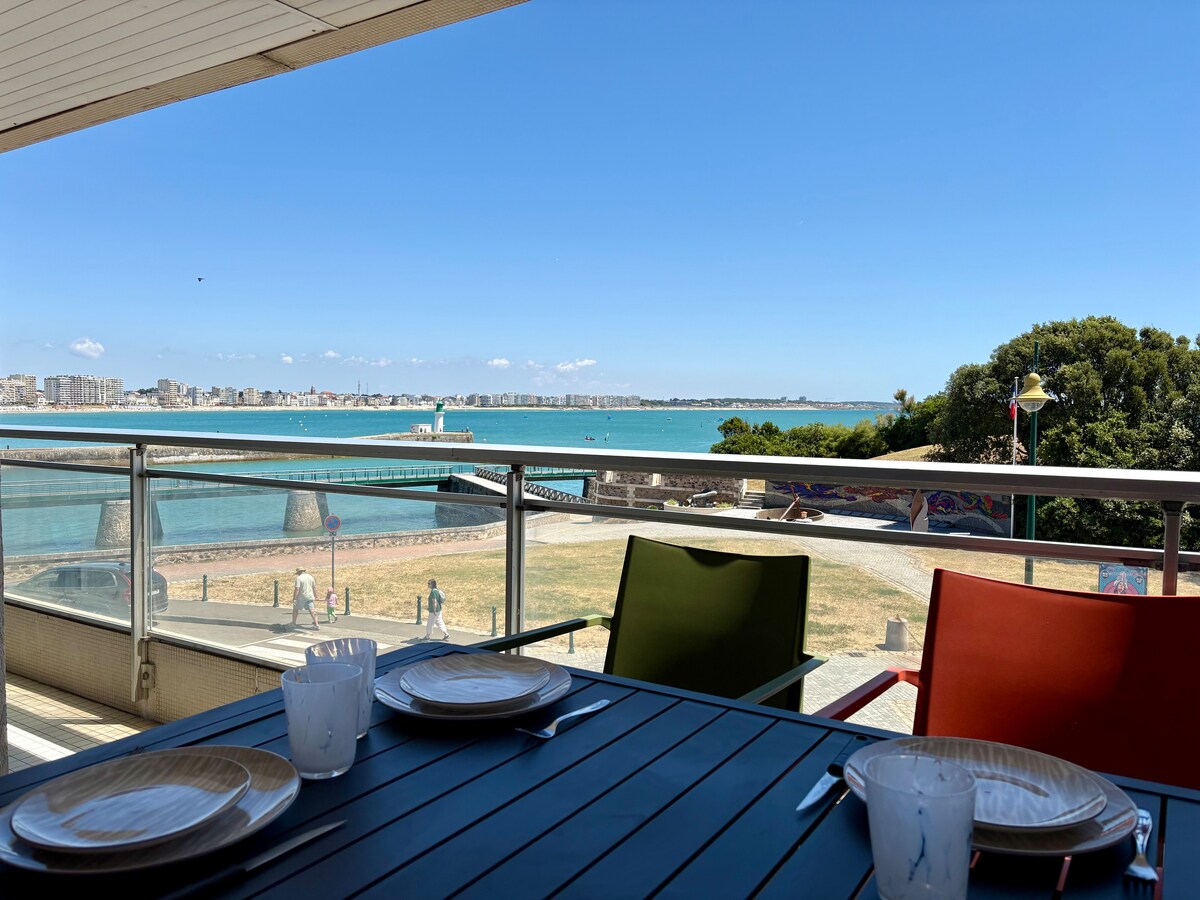 A balcony table is set for dining with plates and glasses, overlooking a panoramic view of the ocean. The vibrant colors of the chairs add a cheerful touch, while the distant shoreline and coastal greenery frame the scene beneath a clear blue sky.