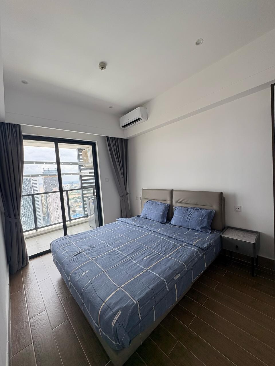 A spacious sleeping area is highlighted by a comfortable bed with blue bedding. A nightstand is positioned beside the bed, and large glass doors open to a balcony, offering views of the surrounding city. Natural light is allowed in through the curtains.