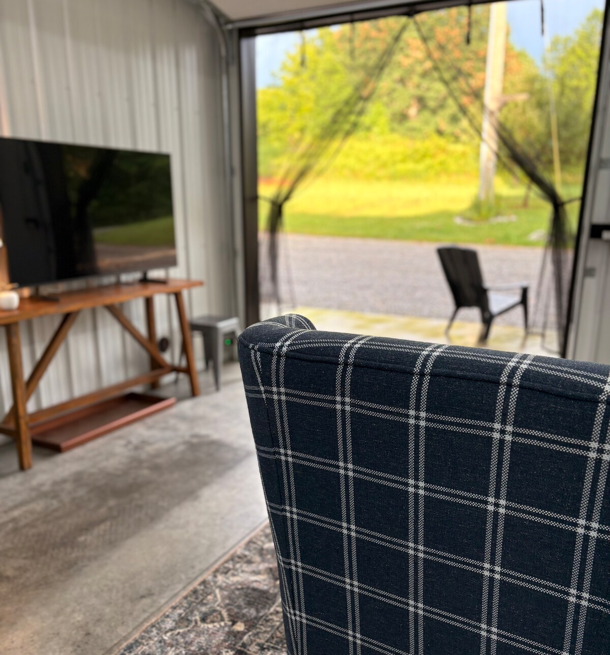A plaid upholstered chair is featured in the foreground, facing a wall-mounted television on a wooden console. The garage door is open, revealing a view of the green outdoors and a chair positioned on the gravel path outside.