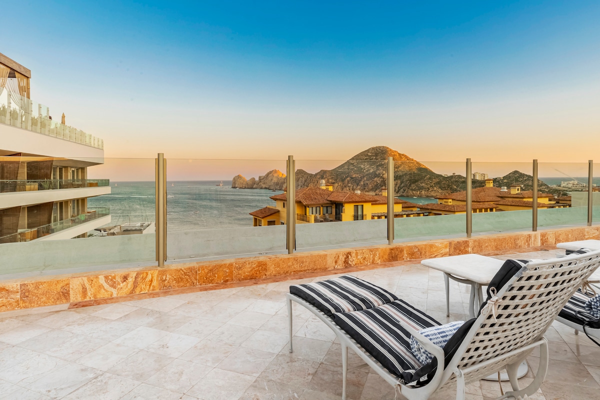 A spacious terrace features comfortable loungers facing stunning ocean views and distant mountains. The area is framed by glass railings, providing safety and unobstructed sightlines. Soft hues from the sunset cast a warm glow over the elegant marble flooring.