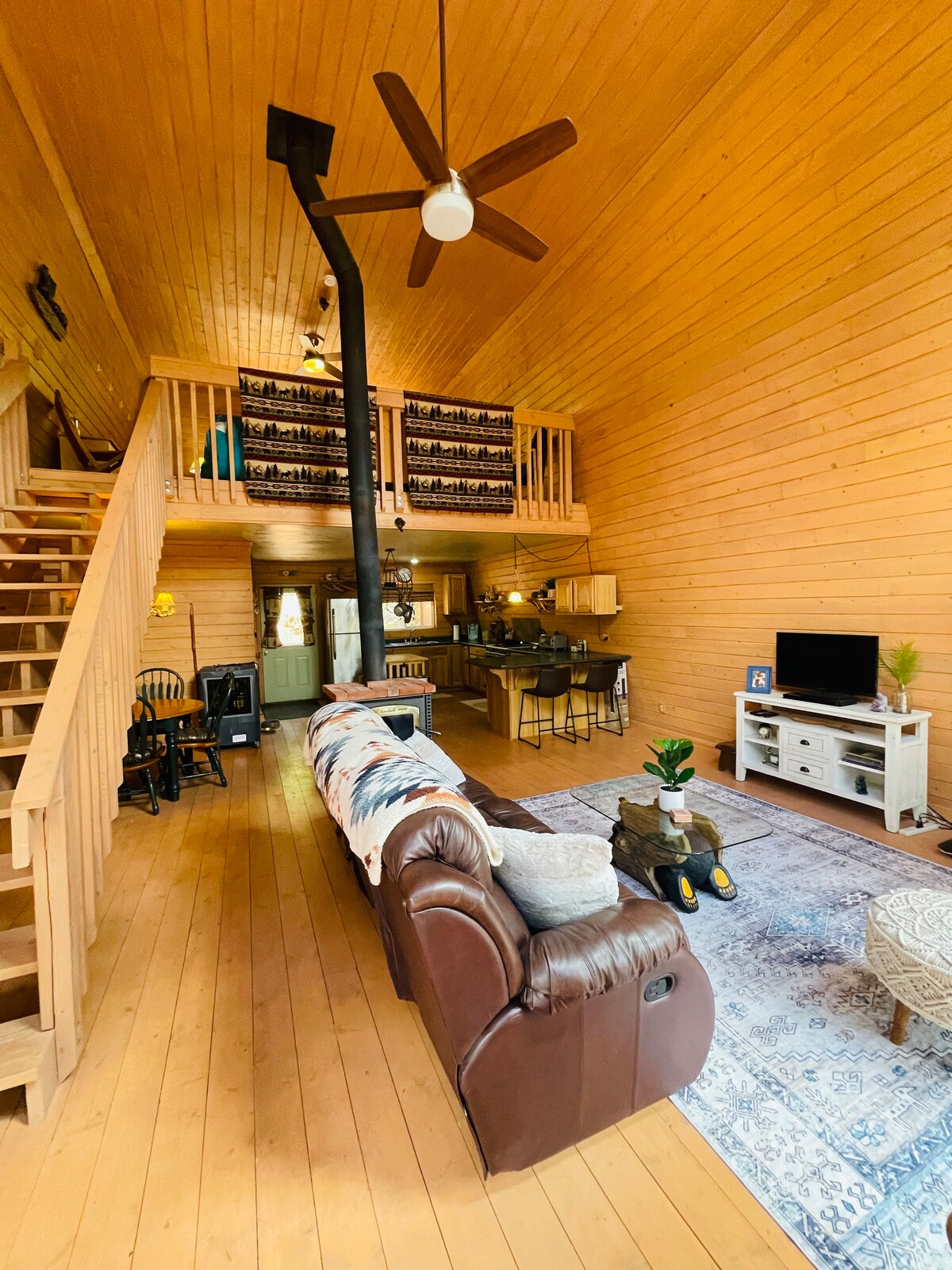 An open-concept living space showcases high wooden ceilings and a staircase leading to a loft. A cozy seating area features a brown leather couch and a decorative rug. A wooden dining table and kitchen area are visible in the background, complemented by a wood-burning stove.