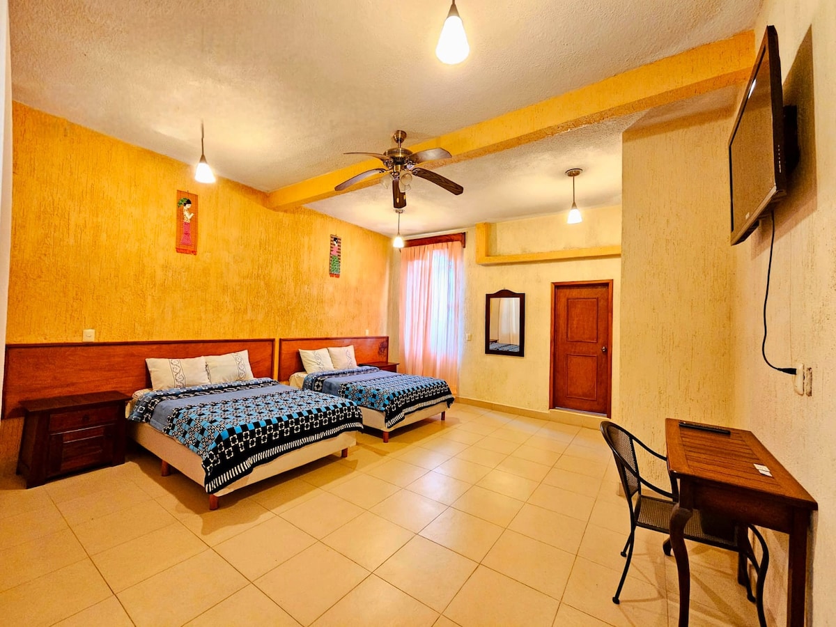 A spacious room is furnished with two double beds framed in wood, accented by bright blue bedding. A small wooden table is positioned near a wall-mounted television. Soft lighting fixtures are suspended from the ceiling, illuminating the warm yellow walls. A mirror and a door are visible.