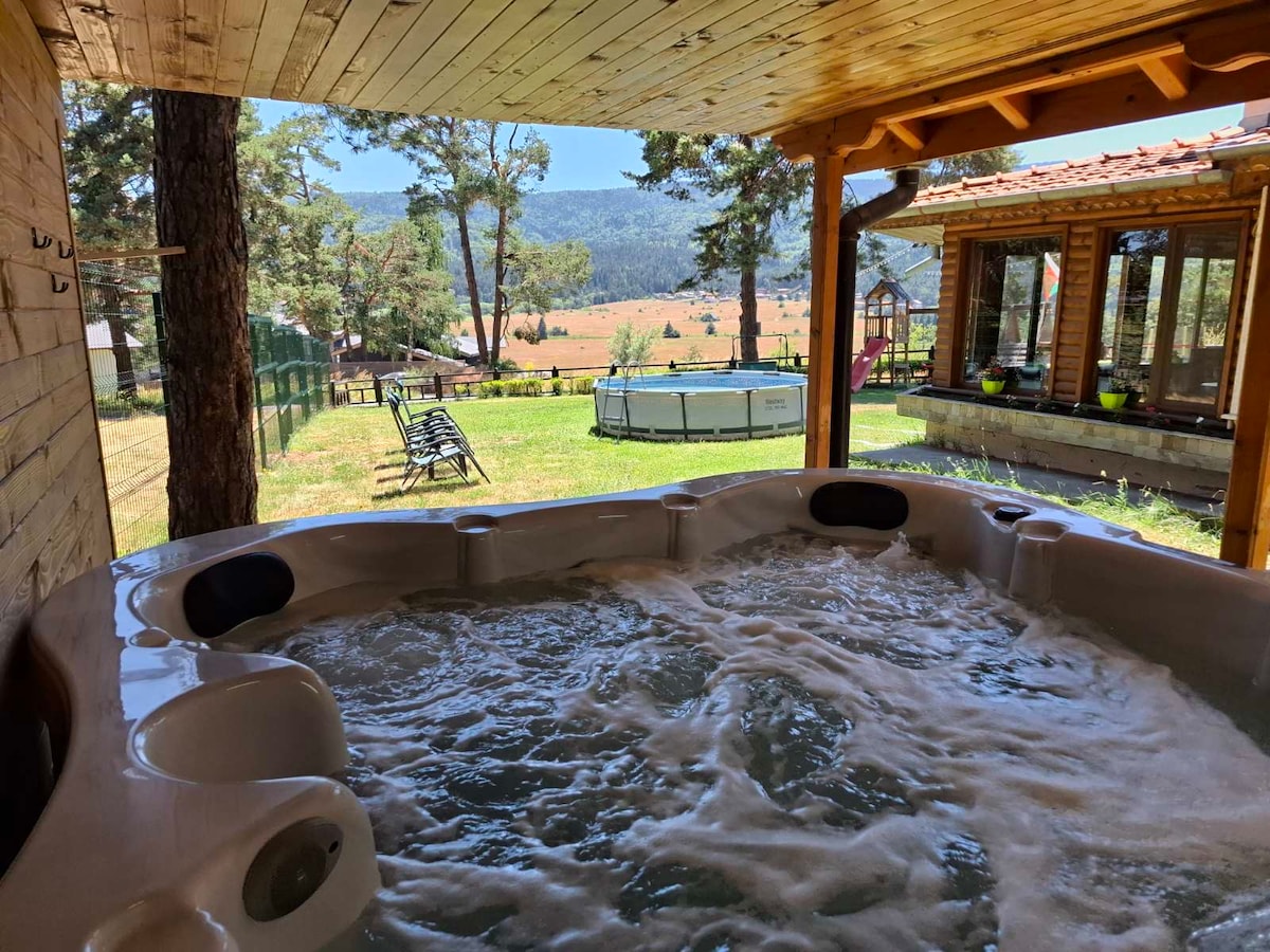 An outdoor eight-person jacuzzi is featured under a wooden pergola, surrounded by lush greenery and mountainous views. Sun loungers are arranged on a grassy area, and a pool is visible in the background, providing a serene setting for relaxation.