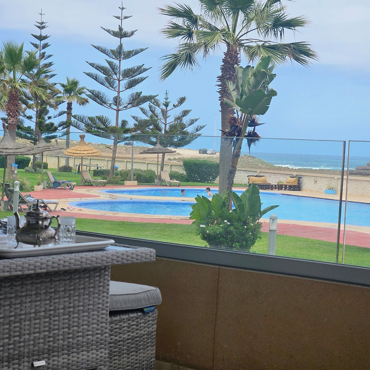A view from the private terrace reveals a tranquil scene with a swimming pool surrounded by lush greenery and palm trees. The ocean is visible in the background, providing a calming atmosphere. Patio furniture, including a wicker table and chairs, is arranged for relaxation.