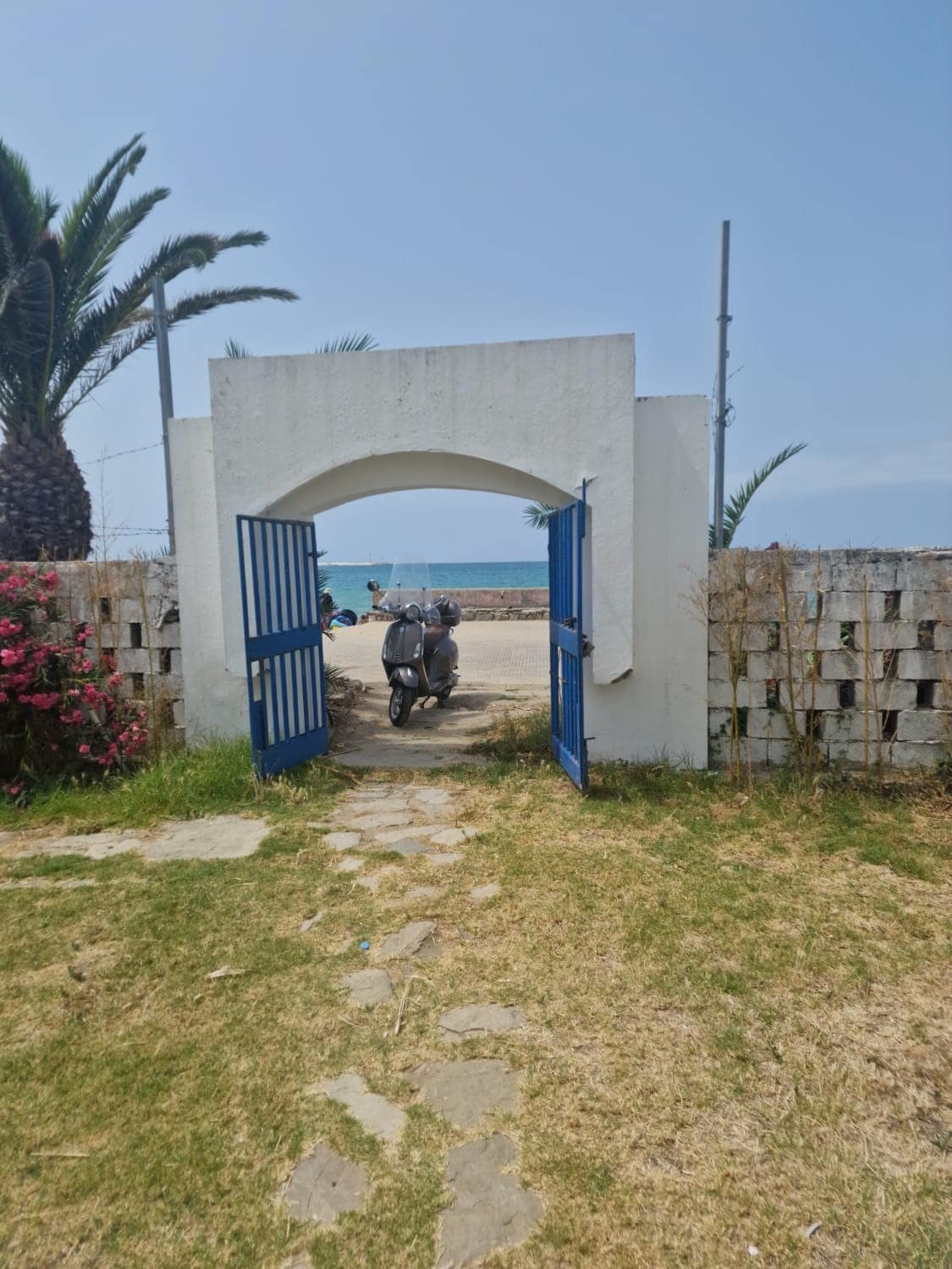 An arched entryway is framed by white walls and vibrant greenery, leading to a sandy pathway. The entrance features double blue gates that open towards a view of the beach and sea in the distance, creating a connection to the outdoor environment.