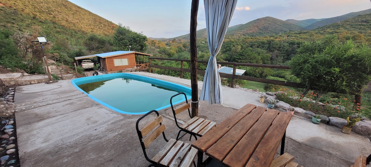A serene outdoor space is featured, showcasing a blue pool set against a backdrop of green hills. A wooden table and chairs are positioned nearby, inviting relaxation. A gazebo with a light curtain offers shade, enhancing the tranquil environment of the natural surroundings.