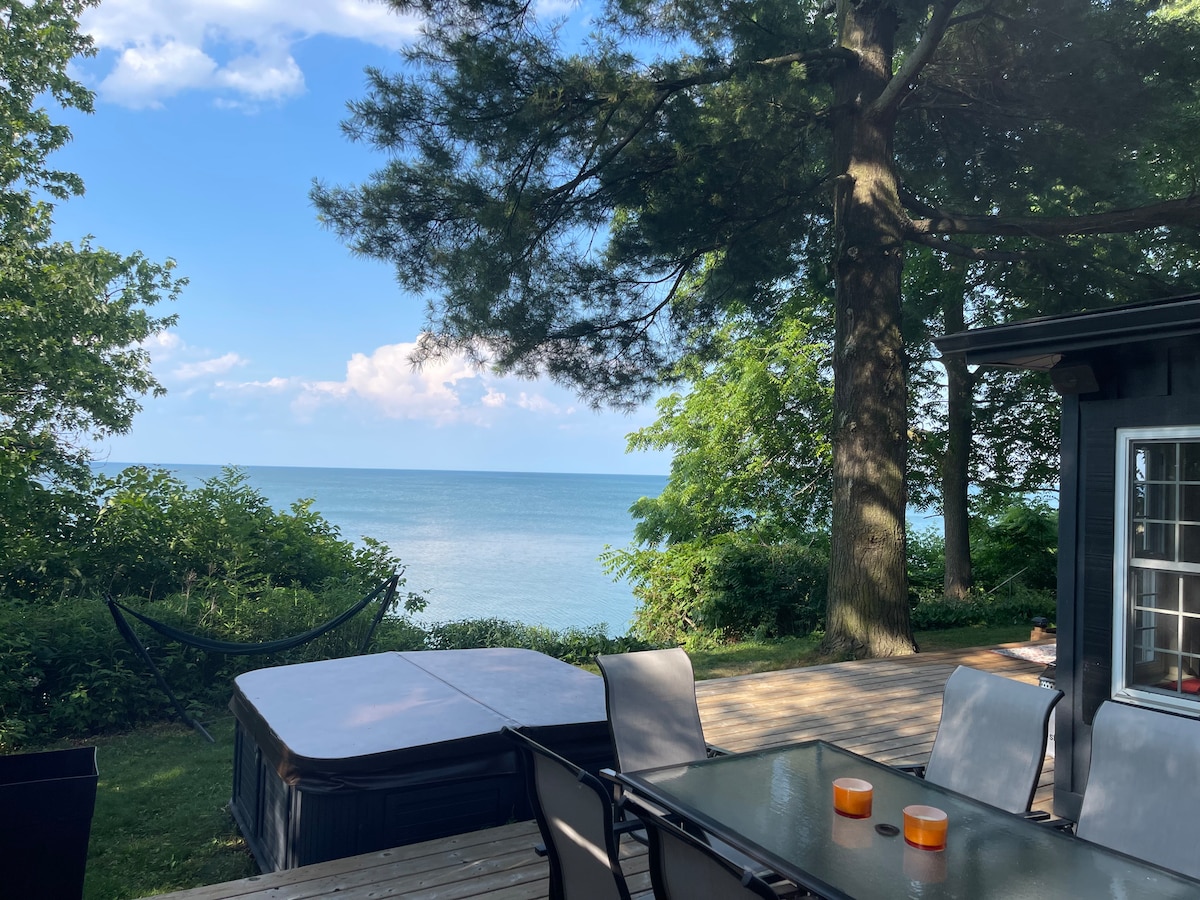 A view of Lake Ontario from the deck, with a private saltwater hot tub positioned nearby. Outdoor seating is provided at a glass-top table, and bright orange candle holders rest on the table. Lush greenery and a hammock are visible alongside the lake.