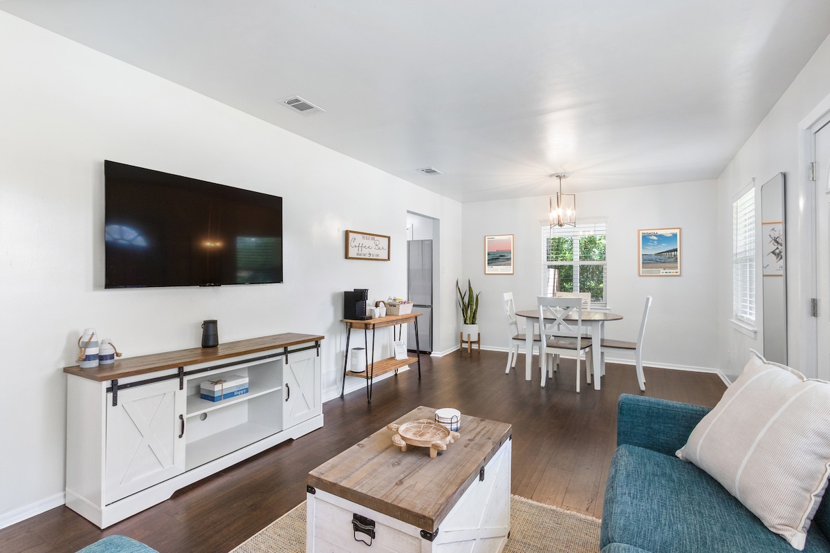A light-filled living area showcases a comfortable seating arrangement with a teal couch and wooden coffee table. A large TV is mounted on the wall. A dining area is visible, featuring a white table surrounded by chairs. Natural light enters through the windows.