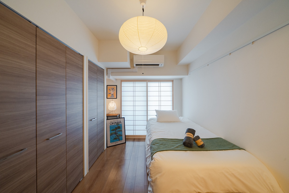 A modern room features a single bed dressed in white linens, complemented by a green throw. A soft light fixture hangs from the ceiling, enhancing the bright atmosphere. Storage options include stylish wood cabinetry, and a decorative piece is displayed against the wall.