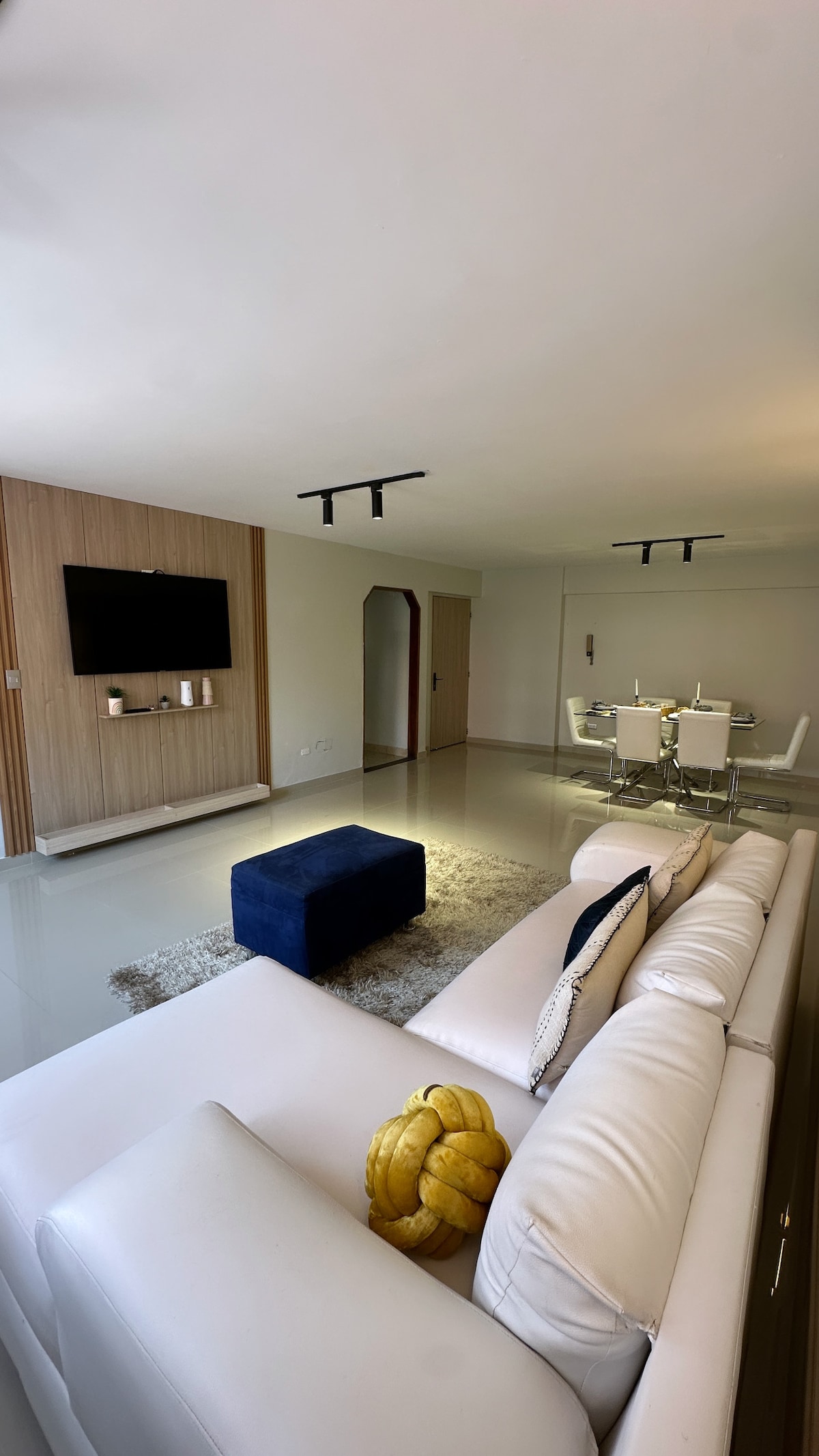 A modern living space features a neutral color palette and sleek furnishings. A white sectional sofa is complemented by a textured rug and a navy ottoman. A wall-mounted television is visible, along with a dining area that includes a glass table and four chairs.