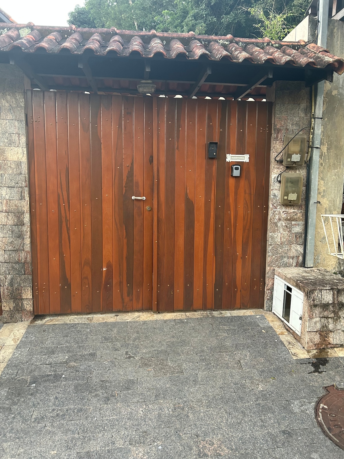 A wooden gate stands at the entrance, featuring vertical slats and a decorative overhang. Stone paving leads up to the gate, offering a clear pathway. Mailboxes are mounted on the adjacent wall, providing functionality to the entrance area.