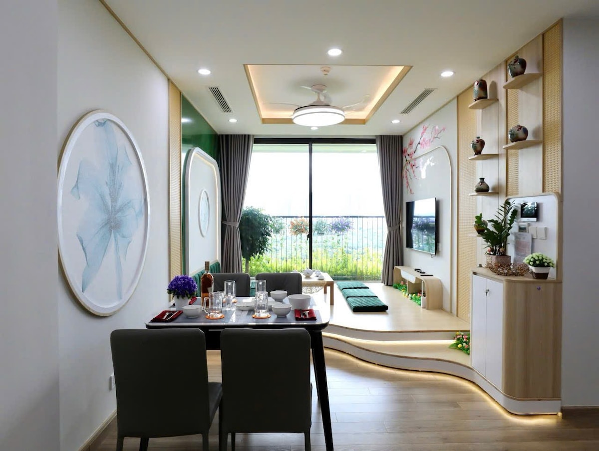 A modern living area is displayed with light-toned wooden flooring and contemporary furniture. A dining table is set for four, while a large window allows natural light to illuminate the space, showcasing green views beyond. Decorative plants and artwork enhance the inviting ambiance.