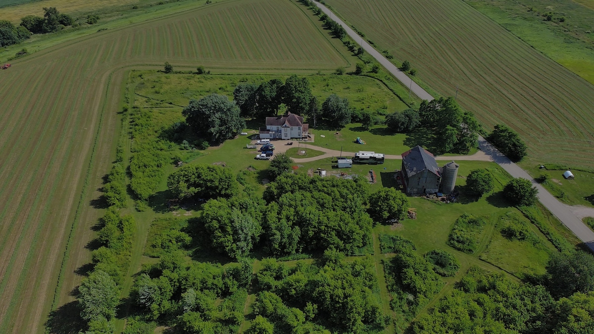 An aerial view showcases an 8-acre farm surrounded by open fields and greenery. The main house is centrally positioned, with several outbuildings nearby. Groomed trails meander through the lush landscape, providing natural pathways for exploration and relaxation.