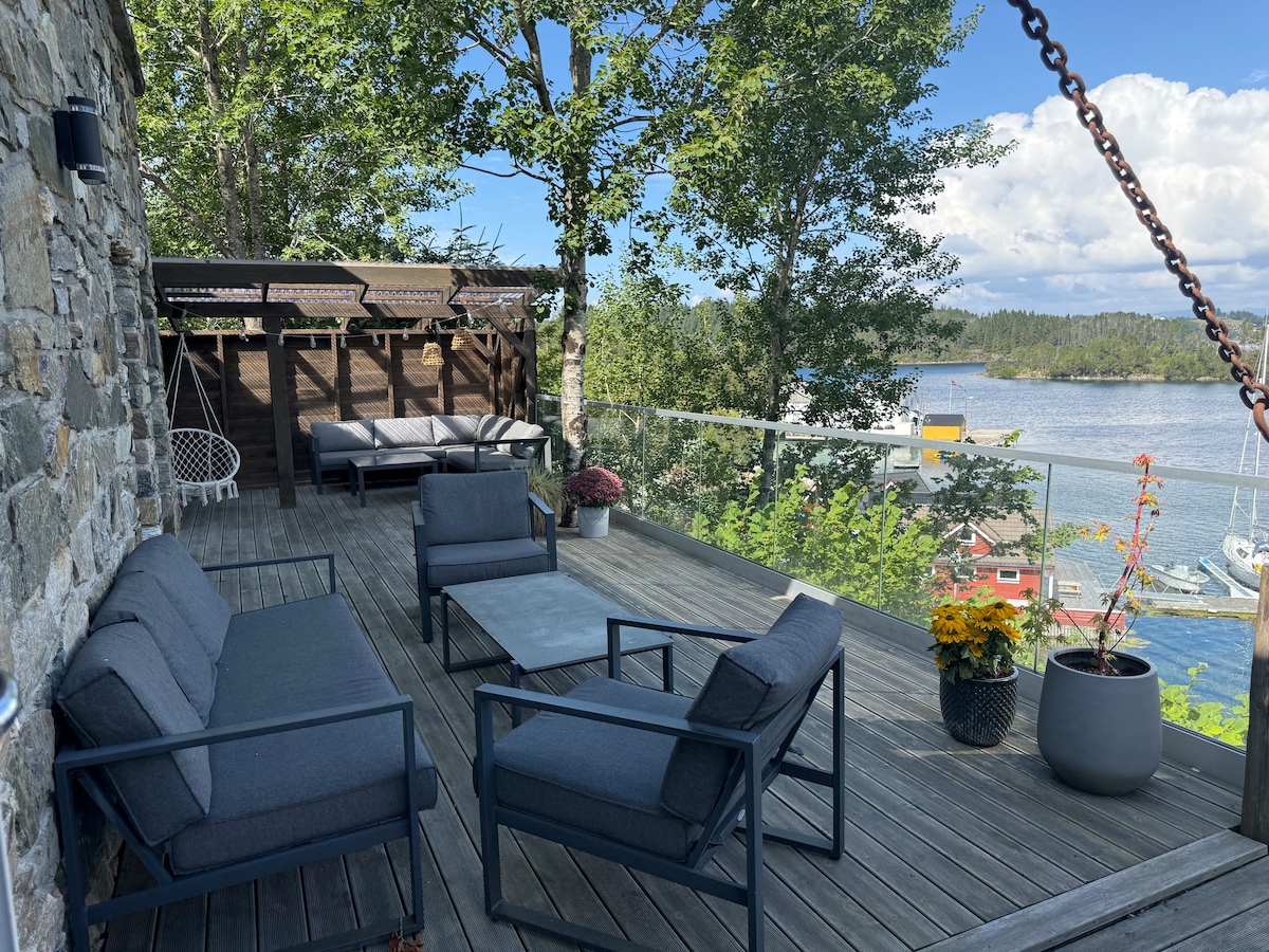 A spacious outdoor terrace features comfortable seating arrangements, including two sofas and multiple chairs, set around a central table. Lush greenery surrounds the area, with panoramic views of the water and boats visible in the distance, creating a calm and inviting atmosphere.